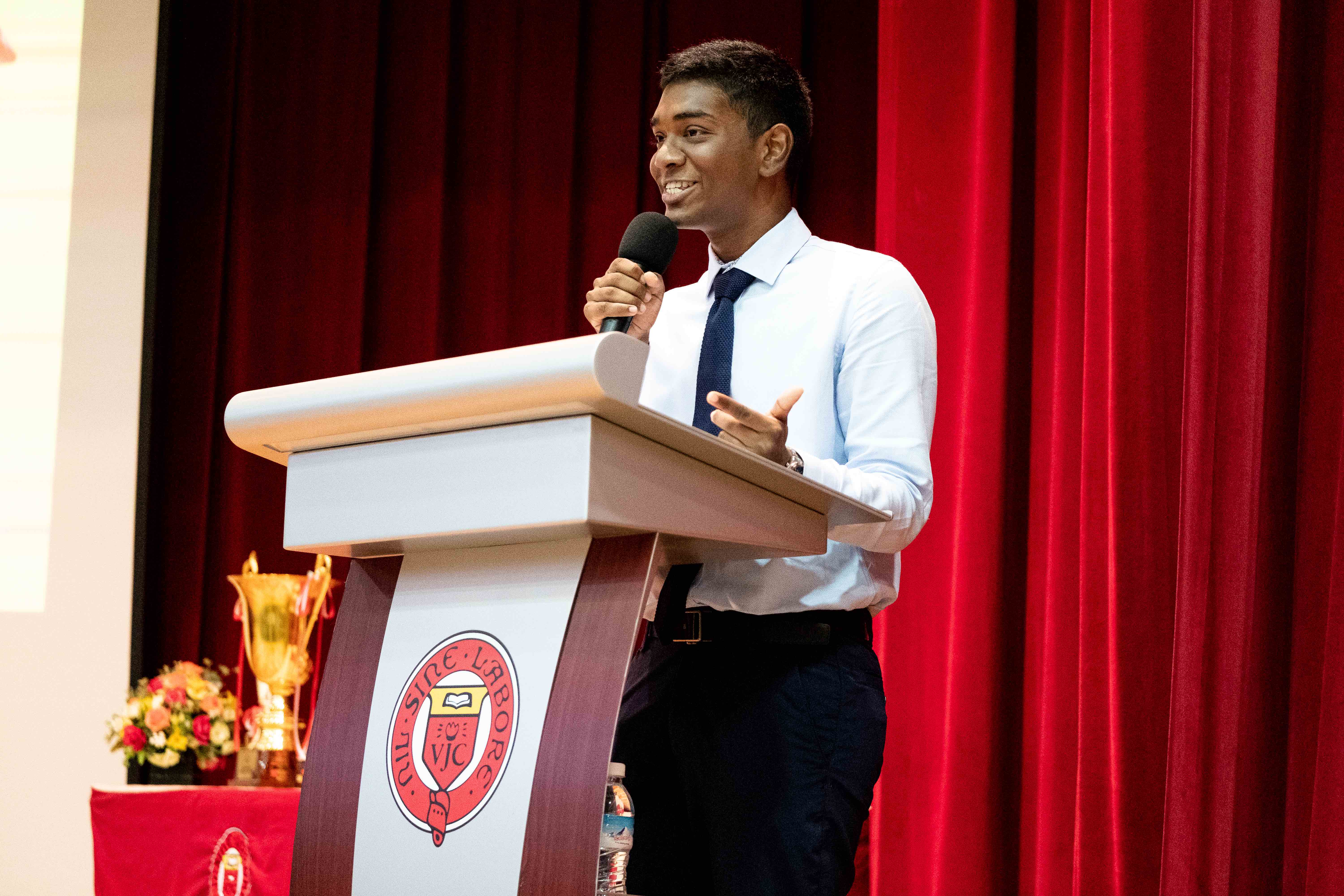 Young man in white shirt and tie smiling while speaking into a microphone at a podium with a school crest.
